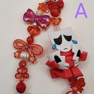 Puede incluir: Un llavero decorativo con un anillo a rayas rojas y blancas, una figura roja con una máscara blanca y lágrimas azules, y una variedad de cuentas rojas, naranjas y transparentes con formas de lazos, corazones, flores y esferas, marcado con la letra A.