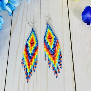 Puede incluir: Un par de pendientes largos de cuentas con un patrón geométrico en tonos de azul, amarillo, naranja y rojo. Los pendientes están hechos de pequeñas cuentas de colores y tienen un diseño de flecos.
