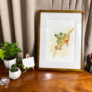 May include: A framed watercolour painting of a green tree frog sitting on a branch. The frog has a light green body with darker green markings and orange feet. The background is a soft yellow watercolour wash.