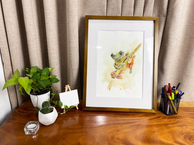 May include: A framed watercolour painting of a green tree frog sitting on a branch. The frog has a light green body with darker green markings and orange feet. The background is a soft yellow watercolour wash.