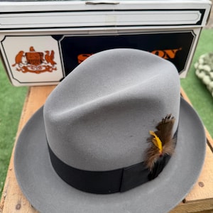 May include: A light gray fedora hat with a black grosgrain ribbon band and a decorative brown and yellow feather accent. The hat is positioned in front of a white and navy blue Stetson Hats cardboard box with a crest logo printed on the front.