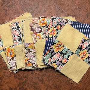 May include: A collection of square fabric patches featuring a mix of solid yellow squares and squares with floral patterns in black, orange, pink, and green, alongside some blue and white striped fabric pieces with frayed edges.