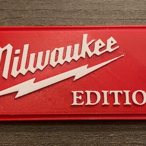 May include: A red rectangular sign featuring the white text "Milwaukee" and "EDITION". A white lightning bolt design is beneath the word "Milwaukee". The sign is likely a product label or emblem.