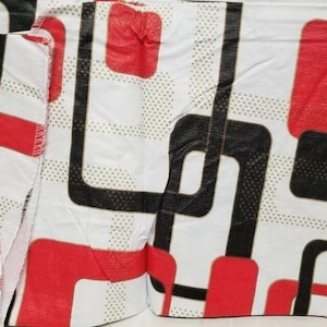 Puede incluir: Primer plano de una tela blanca con un patrón geométrico repetitivo. El diseño presenta formas negras y rojas, incluyendo cuadrados y líneas curvas, sobre un fondo de pequeños puntos dorados. La tela parece estar doblada.