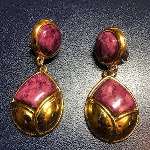 May include: A pair of gold tone dangle earrings with a pink marbled stone. The earrings have a teardrop shape with a square stone in the center.