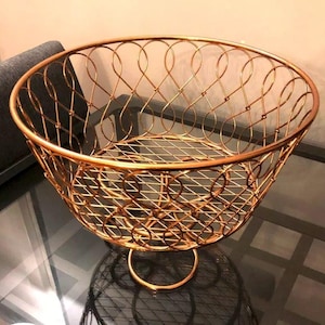 May include: A gold metal fruit bowl with a decorative, open-weave design. The bowl has a round base and a wide, open top.