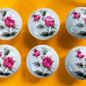 May include: Six white ceramic cabinet knobs with a floral rose design. Each knob features a pink rose with green leaves and stems. The knobs are round and sit against a yellow background.