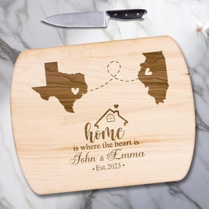May include: A wooden cutting board with a house design and the text "home is where the heart is John & Emma Est. 2023". The board also features outlines of the states of Texas and Illinois with hearts in each state.
