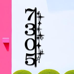 May include: Black metal address sign displaying the numbers 7305. The numbers are vertically aligned on a black bar, with starburst designs on the right side. The sign is mounted on a white wall, with a pink wall and green bushes in the background.