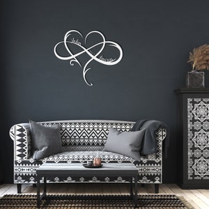 May include: A white metal infinity symbol with a heart in the center. The names "John" and "Lauren" are inscribed on the symbol. The symbol is mounted on a dark gray wall above a black and white patterned sofa.