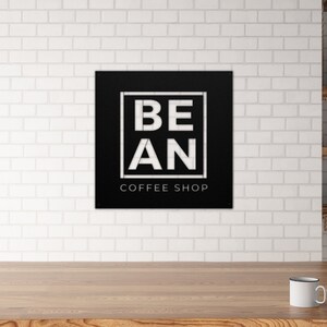 K&ouml;nnte beinhalten: Schwarzes und wei&szlig;es Metallschild mit dem Text "BE AN COFFEE SHOP" in einem wei&szlig;en Quadrat auf schwarzem Hintergrund.