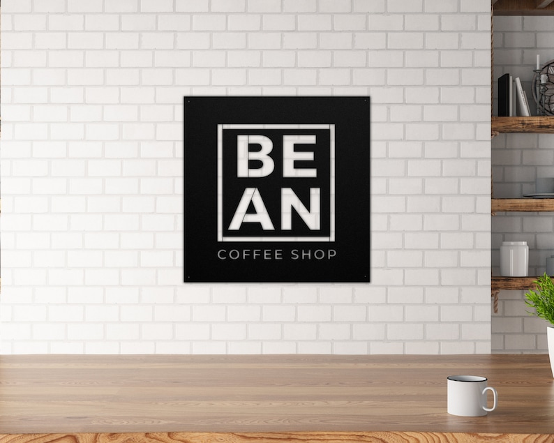 K&ouml;nnte beinhalten: Schwarzes und wei&szlig;es Metallschild mit dem Text "BE AN COFFEE SHOP" in einem wei&szlig;en Quadrat auf schwarzem Hintergrund.