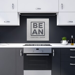 K&ouml;nnte beinhalten: Ein schwarz-wei&szlig;es Schild mit dem Text "BE AN COFFEE SHOP" auf grauem Hintergrund. Das Schild ist an einer schwarzen Wand &uuml;ber einem Edelstahlherd und Backofen in einer K&uuml;che angebracht.