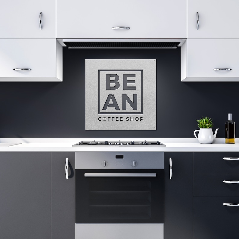 K&ouml;nnte beinhalten: Ein schwarz-wei&szlig;es Schild mit dem Text "BE AN COFFEE SHOP" auf grauem Hintergrund. Das Schild ist an einer schwarzen Wand &uuml;ber einem Edelstahlherd und Backofen in einer K&uuml;che angebracht.