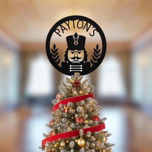 May include: A black metal Christmas tree topper featuring a nutcracker design within a circular frame. The text "PAYTON'S" is arched around the top. The topper is placed on a decorated Christmas tree with red ribbon and ornaments.