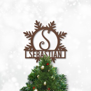 Op de afbeelding: Een bruine metalen kerstboomtopper in de vorm van een sneeuwvlok met de letter "S" in een cirkel en de naam "SEBASTIAN" eronder. De topper staat bovenop een groene kerstboom.