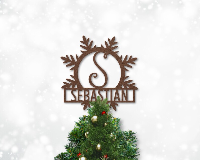 Op de afbeelding: Een bruine metalen kerstboomtopper in de vorm van een sneeuwvlok met de letter "S" in een cirkel en de naam "SEBASTIAN" eronder. De topper staat bovenop een groene kerstboom.
