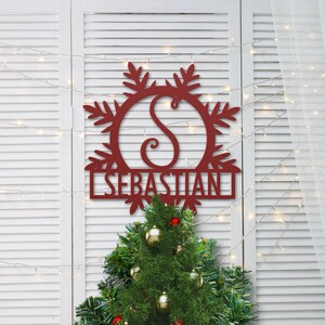 Op de afbeelding: Rode metalen sneeuwvlok met de letter "S" in het midden en de naam "SEBASTIAN" eronder. De sneeuwvlok hangt aan een witte, lattenwand met een kerstboom op de voorgrond.
