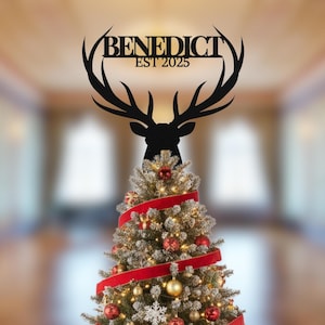 May include: Black metal deer head Christmas tree topper with the name "BENEDICT" and the year "EST 2025". The topper sits atop a decorated Christmas tree with red ribbon and ornaments.