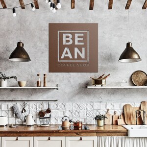 K&ouml;nnte beinhalten: Ein braunes Metallschild mit den Worten "BE AN COFFEE SHOP", die in Wei&szlig; ausgeschnitten sind. Das Schild h&auml;ngt an einer grauen Wand in einer K&uuml;chenumgebung.
