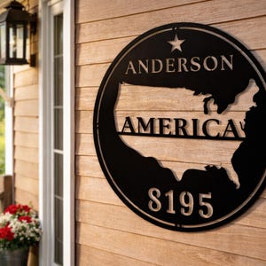 May include: Black metal sign with the name "ANDERSON", the word "AMERICA", and the number "8195" cut out. The sign features a star and the outline of the United States. It is mounted on a wooden wall.