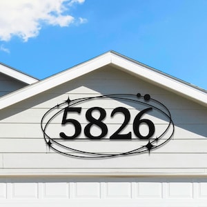 May include: Black metal house number sign with the number 5826 in a stylized font. The numbers are enclosed within an oval design with star accents. The sign is mounted on a light gray house exterior above a white garage door.