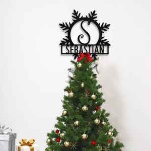 Op de afbeelding: Zwarte metalen sneeuwvlok kerstboomtopper met de initialen "S" en de naam "Sebastian" in een rechthoekige banner.