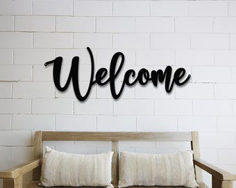 May include: Black metal welcome sign with a cursive font, mounted on a white brick wall above a wooden bench with two beige pillows.
