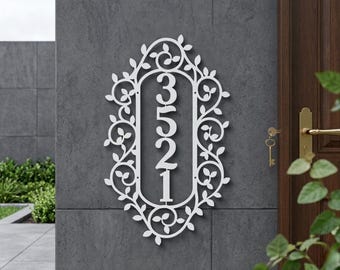 Op de afbeelding: Wit metalen adresbord met de cijfers 3521 in een verticale opstelling. De cijfers staan in een ovale lijst met een decoratief bloemen- en bladerenpatroon. Het bord is bevestigd aan een grijze muur naast een houten deur.