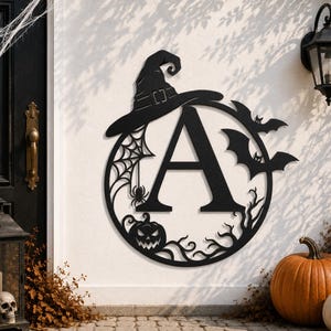 May include: A circular black metal wall sign featuring the letter A in the center. The design includes a witch hat on top, a spider web with a spider on the left, two flying bats on the right, and a jack-o'-lantern pumpkin at the bottom.