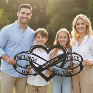 May include: A black metal family name sign featuring intertwined infinity symbols and a heart. The sign displays the names "Melinda", "Mia", "Nick", and "William", along with the date "12.06.2020". The sign is held by a family of four.