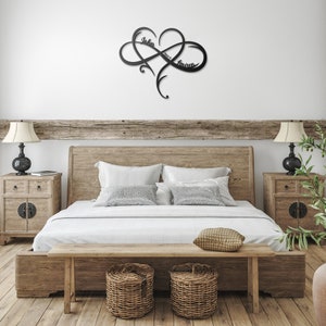 May include: A black metal infinity symbol with a heart in the center. The names "John" and "Lauren" are inscribed on the symbol. The symbol is mounted on a white wall above a wooden bed frame.