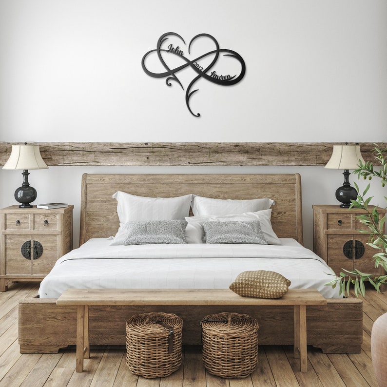 May include: A black metal infinity symbol with a heart in the center. The names "John" and "Lauren" are inscribed on the symbol. The symbol is mounted on a white wall above a wooden bed frame.