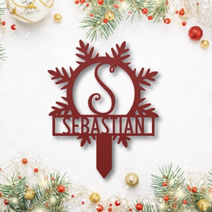 Op de afbeelding: Rode metalen sneeuwvlok ornament met een cursief "S" in het midden en de naam "SEBASTIAN" eronder. Het ornament is ontworpen om te worden gebruikt als een kerstboomtopper.