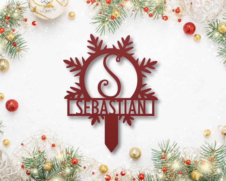 Op de afbeelding: Rode metalen sneeuwvlok ornament met een cursief "S" in het midden en de naam "SEBASTIAN" eronder. Het ornament is ontworpen om te worden gebruikt als een kerstboomtopper.
