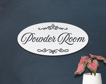 May include: Oval white sign with the words "Powder Room" in elegant black script. The sign has decorative flourishes above and below the text. The sign is mounted on a dark blue wall, with a potted plant in the lower right corner.
