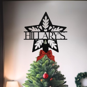 May include: A black metal star Christmas tree topper with the name "Hillary's" cut out. The star has a decorative leaf design and is placed on top of a green Christmas tree with a red bow and ornaments.