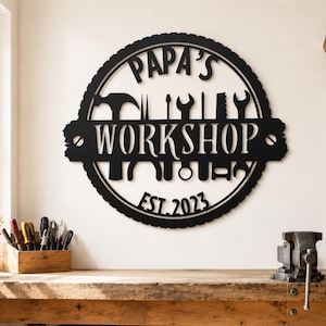 May include: Black metal sign with the words "PAPA'S WORKSHOP EST. 2023" in a circular design. The sign features silhouettes of tools. The sign is mounted on a white wall above a wooden workbench.