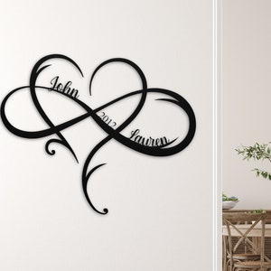 May include: Black metal wall art featuring an infinity symbol intertwined with a heart. The names "John" and "Lauren" and the year "2012" are inscribed within the design.