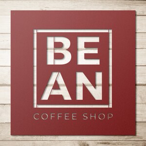 K&ouml;nnte beinhalten: Ein rotes Metallschild mit den Worten "BE AN COFFEE SHOP" in wei&szlig; ausgeschnitten. Das Schild hat einen rustikalen, Vintage-Look.