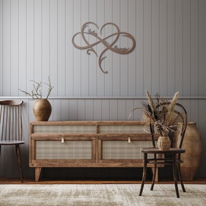 May include: A wooden infinity symbol with the names "John" and "Lauren" and the year "2022" carved into it. The symbol is hanging on a gray wall above a wooden dresser with two drawers. There is a vase with dried branches on the dresser and a chair with a vase of dried flowers next to it.
