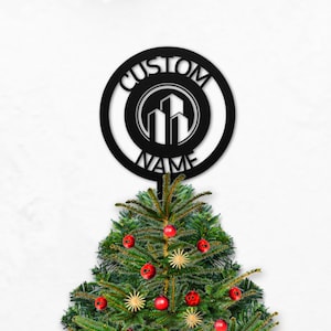 May include: A black metal Christmas tree topper with the words "CUSTOM NAME" encircling a building silhouette. The topper is placed on top of a decorated Christmas tree with red ornaments and star-shaped decorations.