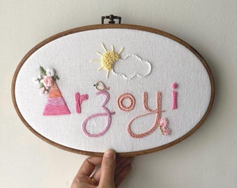 May include: Oval embroidery hoop with the name "Arzoyi" stitched in pink and orange thread. The design includes a sun, cloud, bird, and floral accents. The hoop is held by a hand.