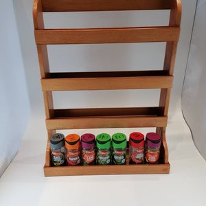 May include: A wooden spice rack with seven glass spice jars. The jars are labelled with the following spices: Black Pepper, Paprika, Lamb, Coriander, Chives, Chilli Cheese, and Fish.