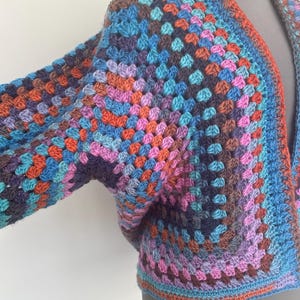 May include: A crochet cardigan featuring a multi-colored granny square pattern. The garment displays a vibrant mix of blue, purple, pink, and orange yarn, constructed with a textured stitch. It has an open front design with wide sleeves.