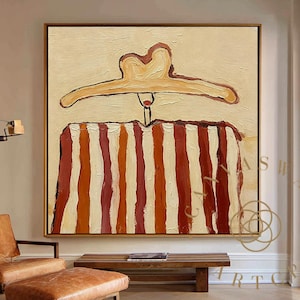 May include: Abstract painting featuring a beige background with a brown and red striped design. A brown coat hanger is at the top. The artwork is framed in a dark brown frame and hangs on a beige wall. The room has a leather chair and ottoman.