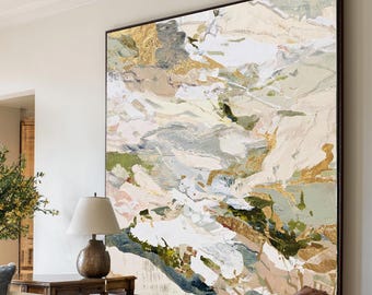 May include: Large abstract painting with a black frame. The artwork features a mix of colors, including gold, white, green, and beige, creating a textured, layered effect. The painting hangs above a brown leather armchair in a living room setting.