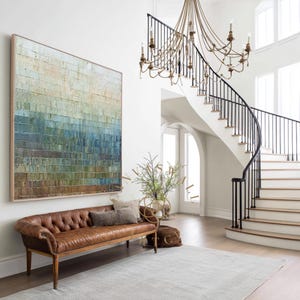 May include: An interior shot of a grand entryway featuring a large abstract painting in shades of blue, green, and brown. A tufted brown leather bench sits below the artwork. A winding staircase with black railings and a chandelier complete the scene.