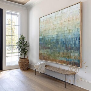 May include: Large abstract painting with a textured surface, featuring horizontal bands of color ranging from beige to blue and green. The artwork is framed in light wood and hangs above a woven bench with black metal legs. A potted plant and a window are also visible.