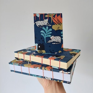 Op de afbeelding: Drie blauwe en witte gepatroneerde dagboeken met rode stiksels. Het bovenste dagboek heeft een ontwerp met luipaarden en planten. Het middelste dagboek heeft een ontwerp met luipaarden en planten. Het onderste dagboek heeft een ontwerp met luipaarden en planten.